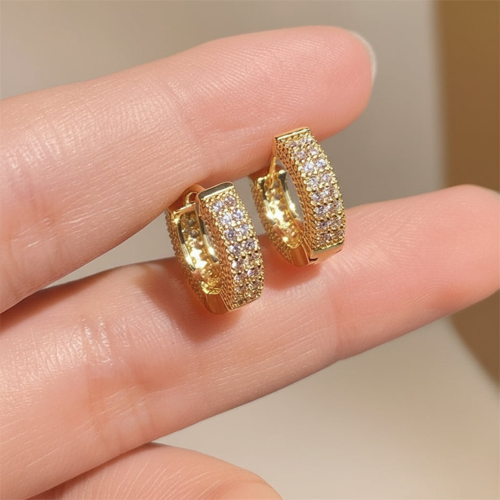 Gold hoop earrings with embedded stones held between fingers against a neutral background